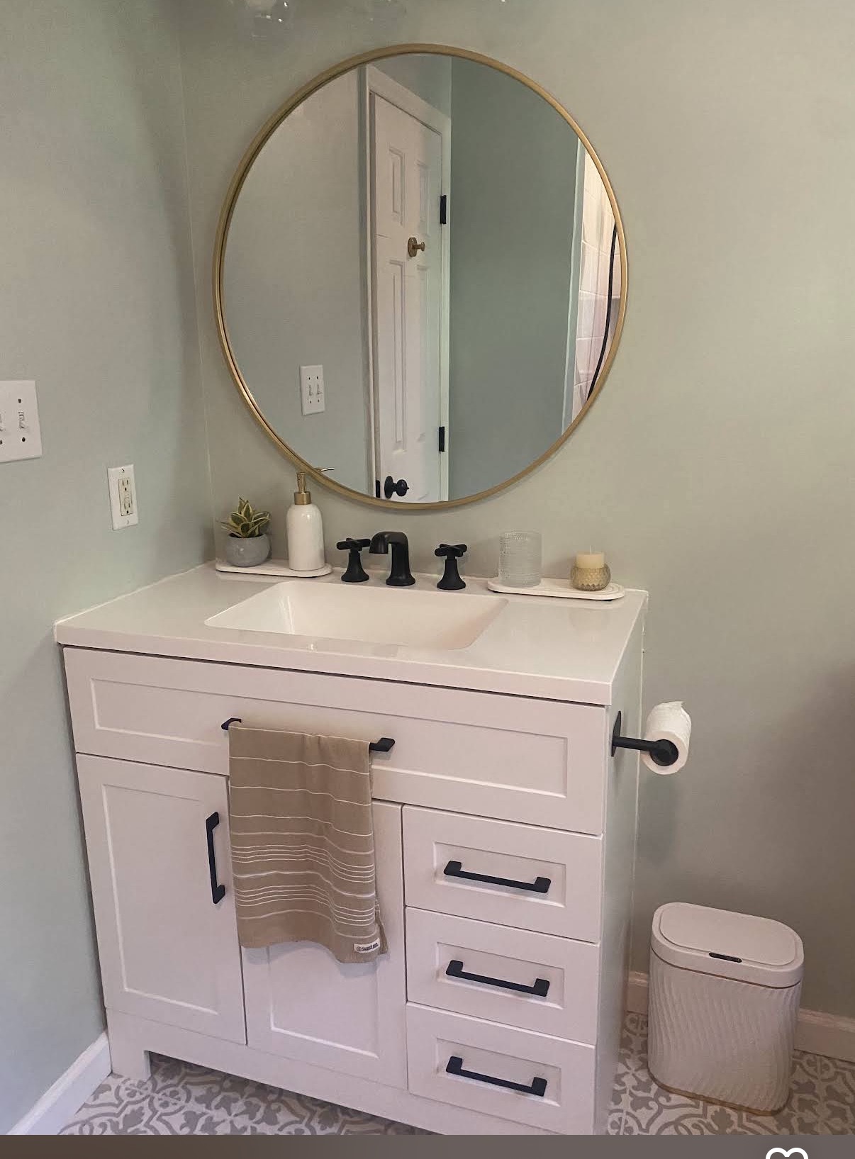 White bathroom vanity with black hardware, round gold-framed mirror and patterned floor